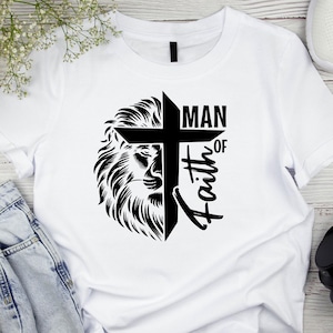 May include: White t-shirt featuring a black graphic design. The design includes a lion's head, a cross, and the words "MAN OF Faith" in a stylized font. The shirt is made of a soft material.