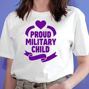 May include: White t-shirt with a purple graphic that reads "PROUD MILITARY CHILD." The design includes a heart, curved lines, and a banner. The shirt is made of a soft material.