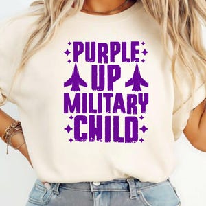 May include: Cream-colored t-shirt with the text "PURPLE UP MILITARY CHILD" in purple. The text includes airplane graphics and starburst accents, creating a patriotic design.