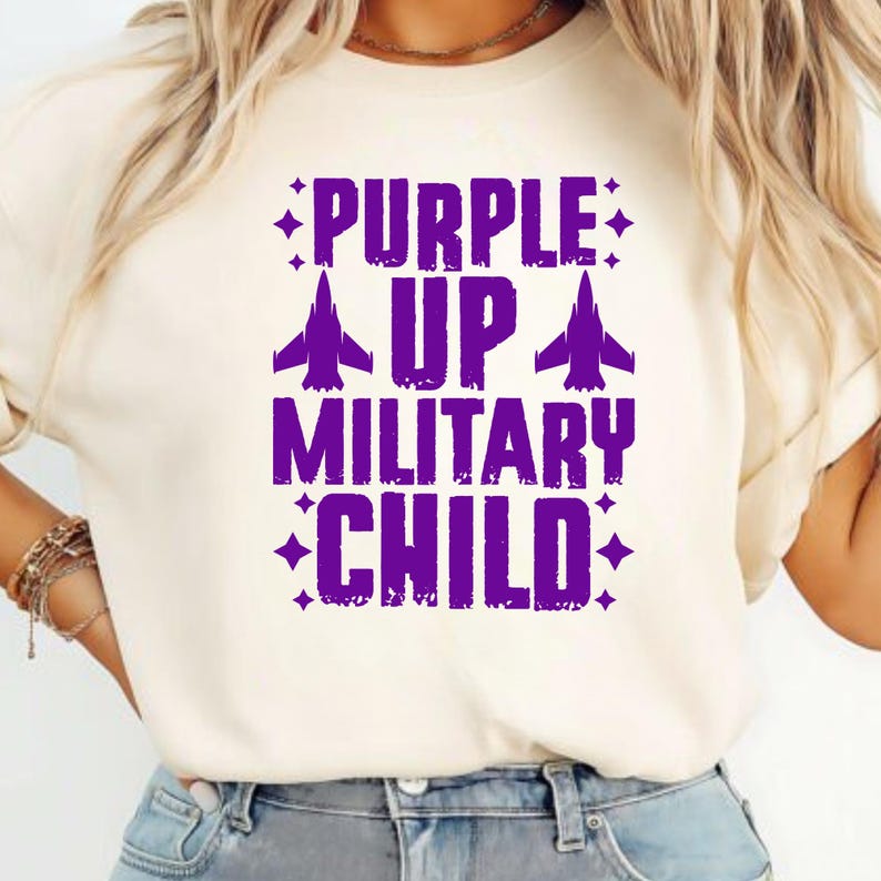 May include: Cream-colored t-shirt with the text "PURPLE UP MILITARY CHILD" in purple. The text includes airplane graphics and starburst accents, creating a patriotic design.