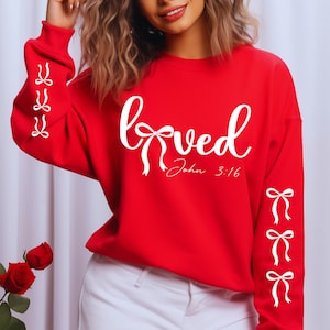 May include: A red sweatshirt with the word "Loved" in white script, featuring a bow design. The text "John 3:16" is below the word "Loved". White bows are also on the sleeves.
