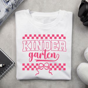 Op de afbeelding: Wit T-shirt met het woord "KINDERGARTEN" in roze, met een geblokt patroon boven en onder de tekst en een strik-afbeelding. Het shirt is gemaakt van een zacht materiaal en is geschikt voor kinderen.