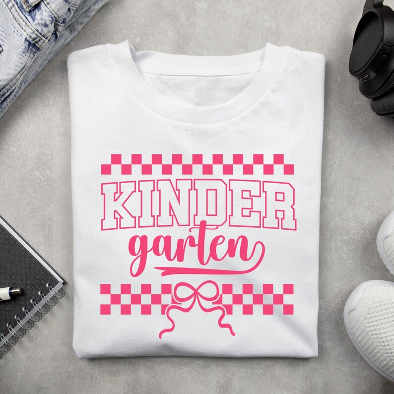 Op de afbeelding: Wit T-shirt met het woord "KINDERGARTEN" in roze, met een geblokt patroon boven en onder de tekst en een strik-afbeelding. Het shirt is gemaakt van een zacht materiaal en is geschikt voor kinderen.
