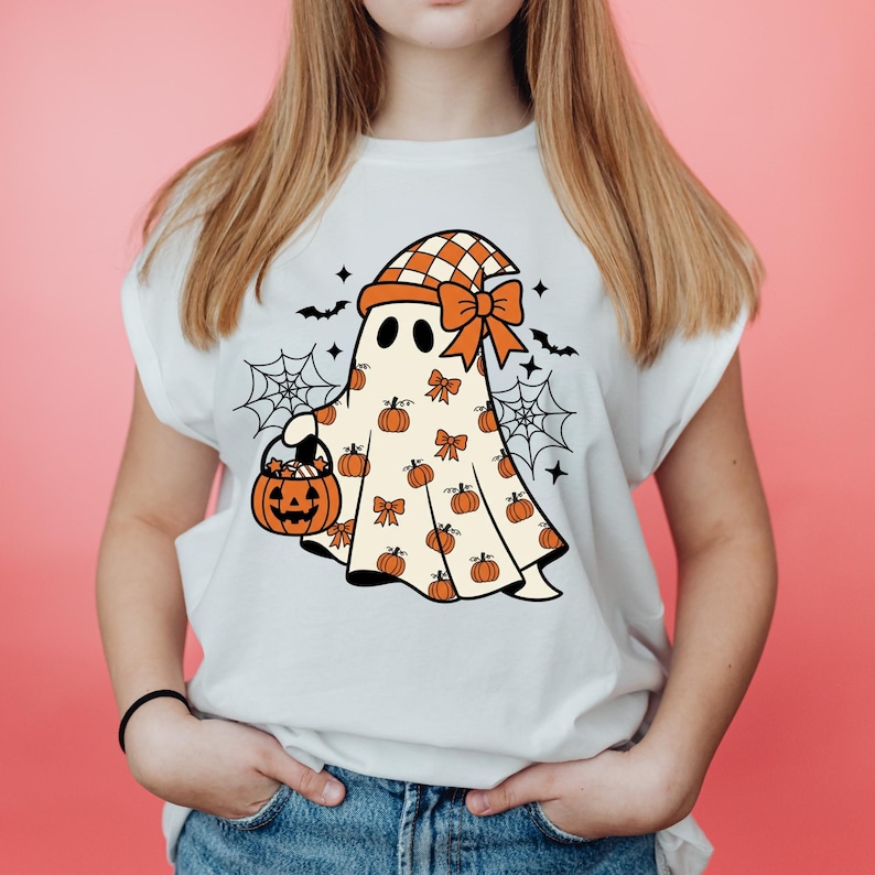 May include: A white t-shirt featuring a graphic of a ghost wearing a checkered hat with an orange bow. The ghost is patterned with small orange pumpkins and bows, holding a jack-o'-lantern bucket, surrounded by black spiderwebs and bats.