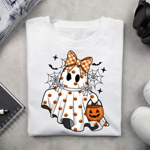 May include: A white t-shirt featuring a graphic of a ghost wearing an orange and white chequered bow. The ghost is covered in small orange pumpkin patterns, holding an orange jack-o'-lantern bucket, surrounded by black spiderwebs and bats.