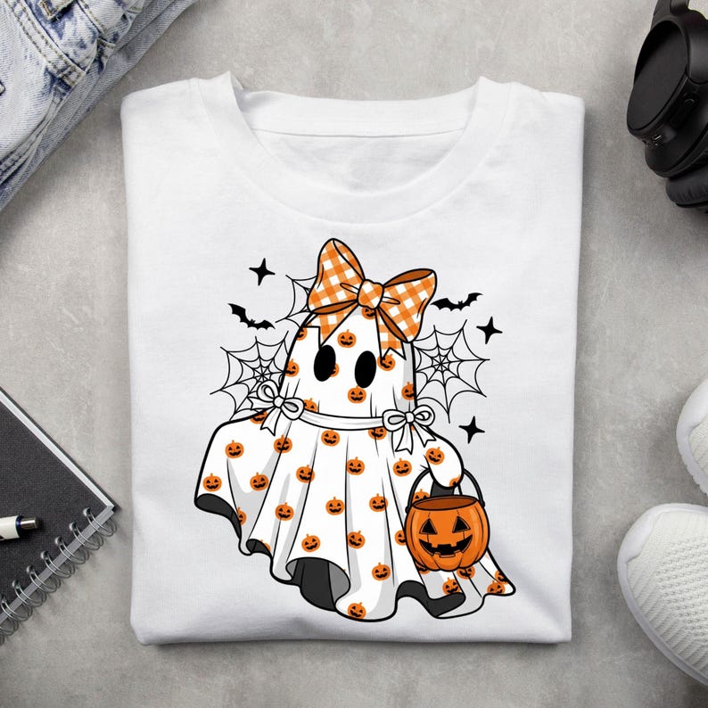 May include: A white t-shirt featuring a graphic of a ghost wearing an orange and white chequered bow. The ghost is covered in small orange pumpkin patterns, holding an orange jack-o'-lantern bucket, surrounded by black spiderwebs and bats.