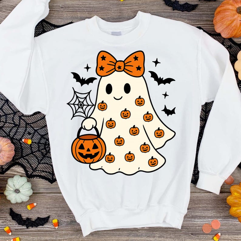 May include: A white long-sleeve sweatshirt featuring a central graphic of a cream-coloured ghost wearing an orange bow with black stars. The ghost is holding an orange jack-o'-lantern bucket and is surrounded by small orange pumpkins, black bats, and a spider web.