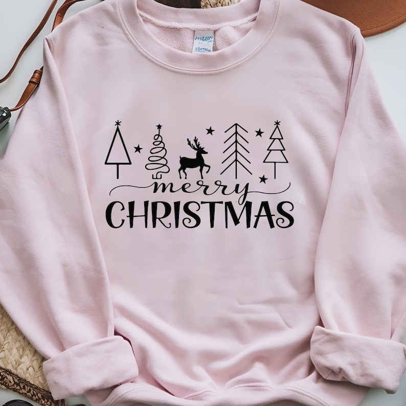 May include: A light pink sweatshirt with a black graphic design. The design includes stylized Christmas trees, a reindeer, stars, and the words "merry CHRISTMAS" in a script font.
