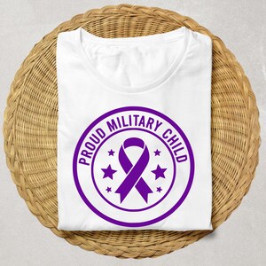 May include: White t-shirt with a crew neck, featuring a purple circular design. The design includes a ribbon and the text "PROUD MILITARY CHILD" in a circular pattern, with stars. The shirt is folded on a woven, light brown surface.
