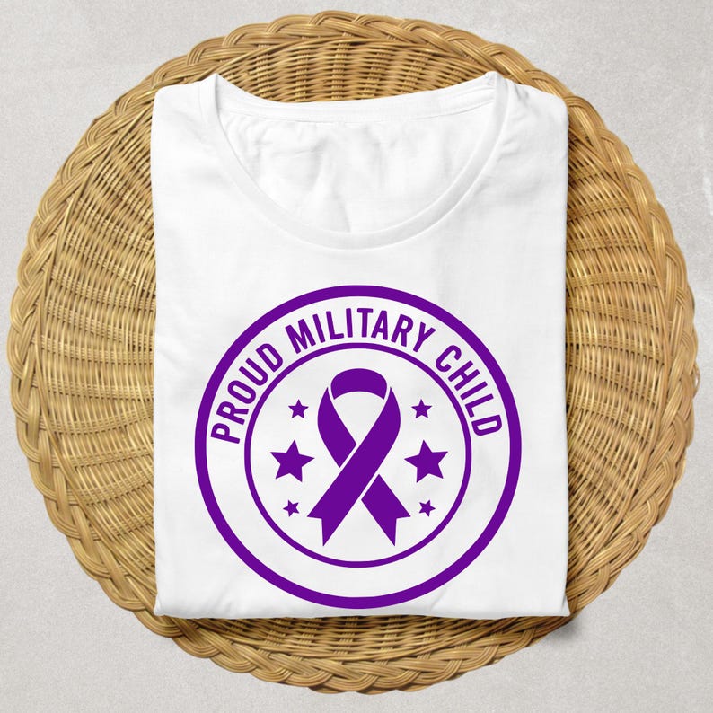 May include: White t-shirt with a crew neck, featuring a purple circular design. The design includes a ribbon and the text "PROUD MILITARY CHILD" in a circular pattern, with stars. The shirt is folded on a woven, light brown surface.