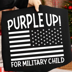 May include: Black t-shirt with a white American flag design. The text "PURPLE UP!" is at the top, and "FOR MILITARY CHILD" is at the bottom. The shirt is held up by a person.