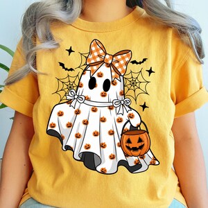 May include: A mustard yellow t-shirt featuring a graphic of a white ghost wearing an orange and white checkered bow. The ghost is covered in small orange pumpkin patterns and holds an orange jack-o'-lantern bucket, surrounded by black spiderwebs.