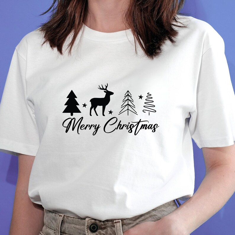 May include: White t-shirt featuring a black graphic of a deer, Christmas trees, and stars, with the text "Merry Christmas" in a script font. The shirt is a classic crew neck style.