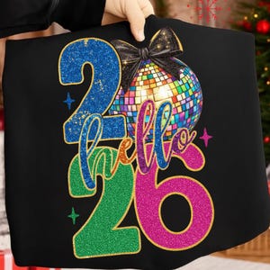 Pode incluir: Camiseta preta com um gráfico de bola de discoteca colorida e o texto "hello 26". Os números "2" e "6" são brilhantes em azul e rosa. Uma fita preta está no topo. O design é festivo.