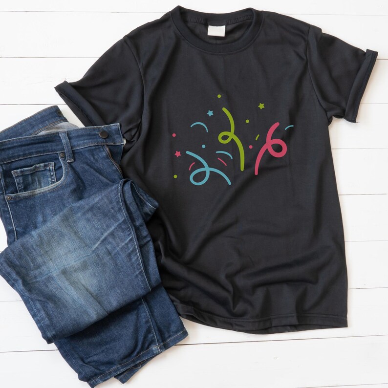 K&ouml;nnte beinhalten: Ein schwarzes T-Shirt mit einem bunten Konfetti-Design. Das Shirt liegt flach auf einer wei&szlig;en Holzoberfl&auml;che mit einer daneben gefalteten blauen Jeans.