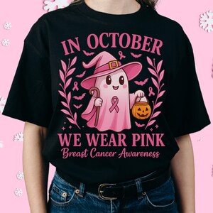 May include: Black t-shirt with a pink graphic featuring a ghost wearing a witch hat, holding a cane and a jack-o'-lantern. The text reads "IN OCTOBER WE WEAR PINK Breast Cancer Awareness". Pink ribbons and bats surround the ghost.
