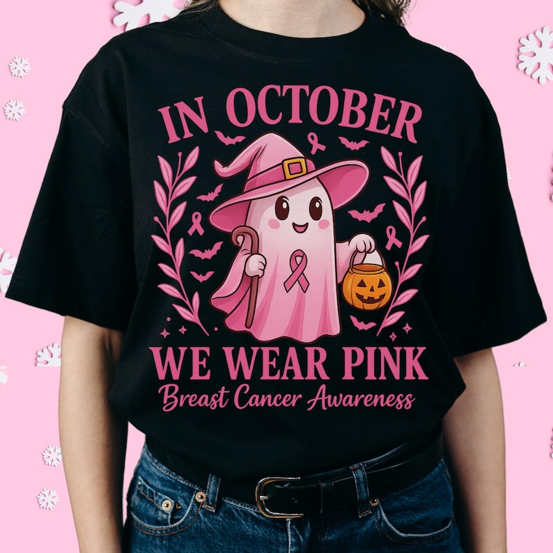 May include: Black t-shirt with a pink graphic featuring a ghost wearing a witch hat, holding a cane and a jack-o'-lantern. The text reads "IN OCTOBER WE WEAR PINK Breast Cancer Awareness". Pink ribbons and bats surround the ghost.