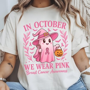 May include: Cream-colored t-shirt with a pink graphic for Breast Cancer Awareness. The design features a friendly ghost wearing a witch hat and holding a cane and a jack-o'-lantern. Text reads "In October We Wear Pink".