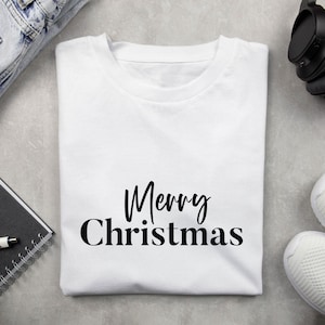 May include: A white t-shirt with the words "Merry Christmas" in black script. The shirt is folded and lies on a gray surface, with a pair of jeans, headphones, and sneakers visible in the background. A notebook and pen are also visible.