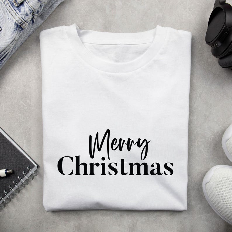 May include: A white t-shirt with the words "Merry Christmas" in black script. The shirt is folded and lies on a gray surface, with a pair of jeans, headphones, and sneakers visible in the background. A notebook and pen are also visible.
