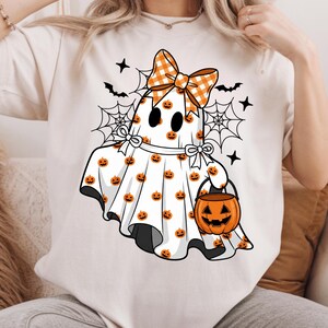 May include: A cream-coloured t-shirt featuring a graphic of a white ghost covered in small orange jack-o'-lanterns. The ghost wears a large orange and white gingham bow, has black eyes, and carries an orange jack-o'-lantern trick-or-treat bucket.