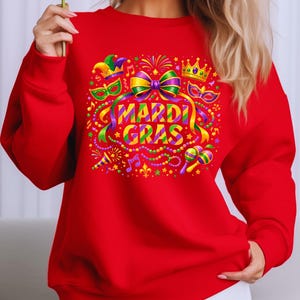 May include: A red sweatshirt featuring a colorful Mardi Gras design. The graphic includes a bow, crown, mask, musical notes, and the words "MARDI GRAS" in a vibrant, multi-colored font. The design is surrounded by festive elements.