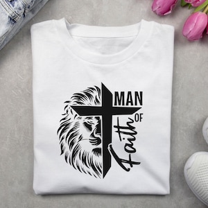 May include: White t-shirt featuring a black graphic design. The design includes a lion's head, a cross, and the words "MAN OF Faith". The shirt is folded and appears to be made of cotton or a similar fabric.