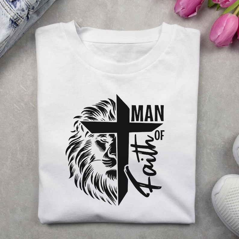 May include: White t-shirt featuring a black graphic design. The design includes a lion's head, a cross, and the words "MAN OF Faith". The shirt is folded and appears to be made of cotton or a similar fabric.