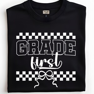 Op de afbeelding: Zwart T-shirt met de witte tekst "GRADE first", omringd door een dambordpatroon. Een witte strik staat onder de tekst. Het T-shirt is opgevouwen.