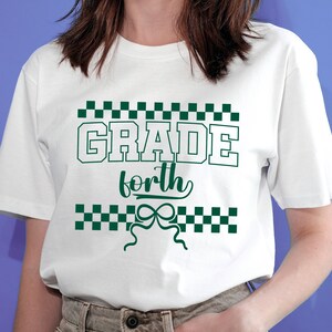 Op de afbeelding: Wit t-shirt met de tekst "GRADE forth" in groene blokletters, omgeven door een groen dambordpatroon. Onder het woord "forth" staat een groen strikdesign.