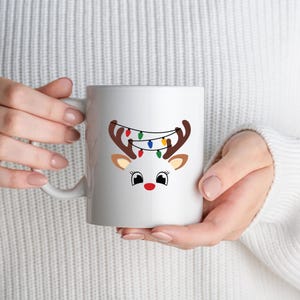 May include: White ceramic mug featuring a reindeer design with brown antlers, tan ears, and a red nose. The reindeer has a string of colorful Christmas lights draped over its antlers. The mug is held by a person wearing a white sweater.
