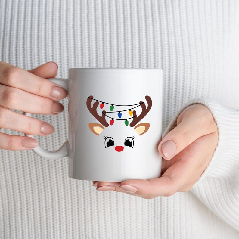 May include: White ceramic mug featuring a reindeer design with brown antlers, tan ears, and a red nose. The reindeer has a string of colorful Christmas lights draped over its antlers. The mug is held by a person wearing a white sweater.