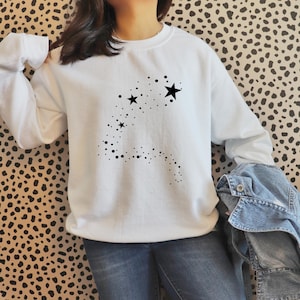 K&ouml;nnte beinhalten: Wei&szlig;er Pullover mit einem schwarzen Stern-Design. Das Design zeigt eine Spur aus kleinen schwarzen Punkten, die zu einem gr&ouml;&szlig;eren Stern f&uuml;hren.
