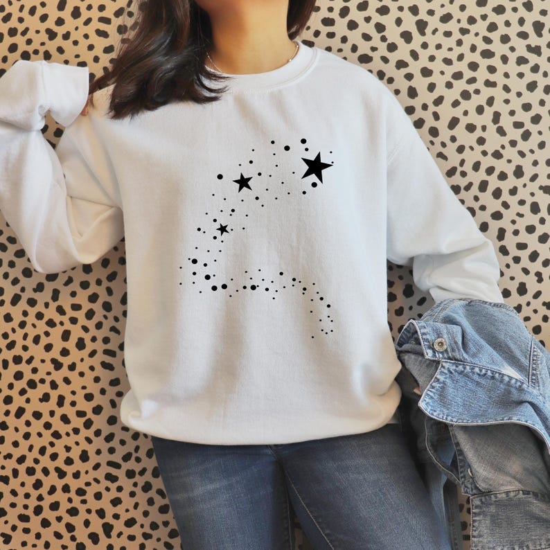 K&ouml;nnte beinhalten: Wei&szlig;er Pullover mit einem schwarzen Stern-Design. Das Design zeigt eine Spur aus kleinen schwarzen Punkten, die zu einem gr&ouml;&szlig;eren Stern f&uuml;hren.