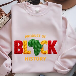 K&ouml;nnte beinhalten: Hellrosa Sweatshirt mit dem Text "PRODUCT OF BLACK HISTORY". Das Wort "BLACK" ist in einem strukturierten Design in Rot, Gelb und Gr&uuml;n gehalten, mit der Form Afrikas in Gr&uuml;n.