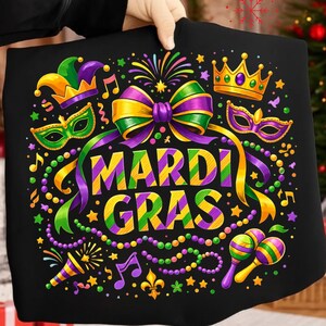May include: Black graphic tee with a vibrant Mardi Gras design. Features a large bow, crown, mask, and maracas in purple, green, and gold. The words "Mardi Gras" are prominently displayed in a festive font, surrounded by stars and musical notes.