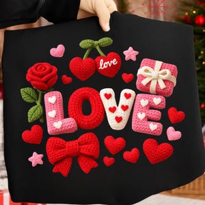 May include: A black fabric with a crochet design featuring the word "LOVE" in red, white, and pink. The design includes hearts, a rose, a bow, a gift box, and stars. The overall theme is romance and Valentine's Day.