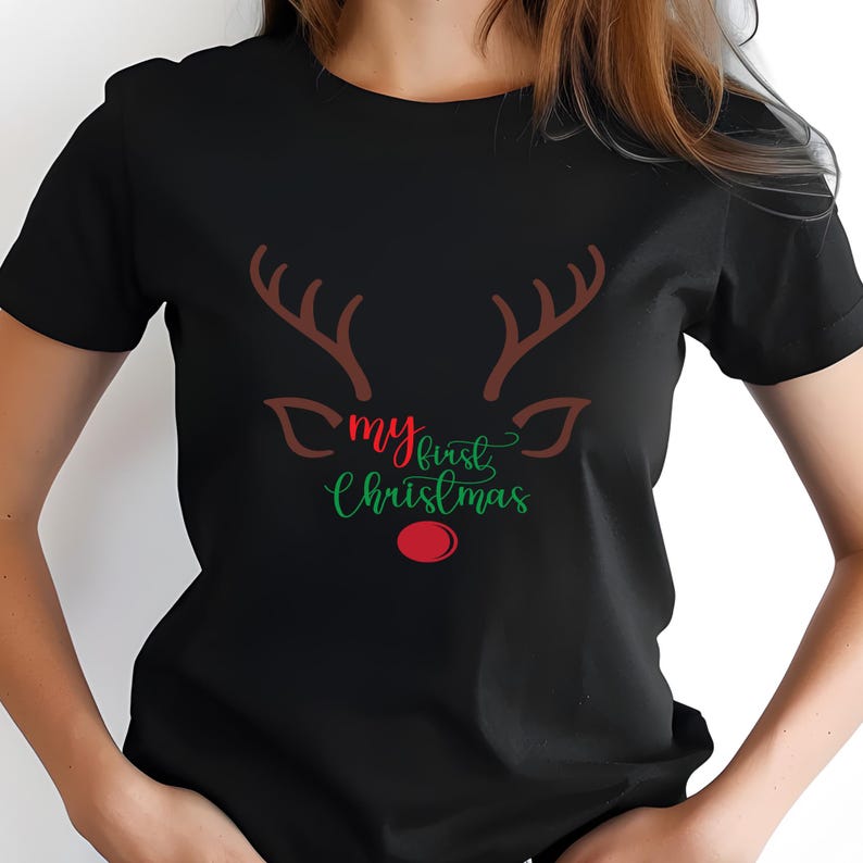 May include: Black t-shirt featuring a reindeer face design. The design includes brown antlers, a red nose, and the text "my first Christmas" in green and red script. A festive holiday-themed tee.