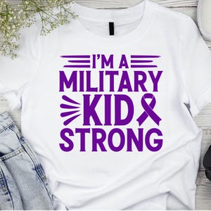 May include: White t-shirt with the purple text "I'M A MILITARY KID STRONG." The design includes a ribbon and stylized text. The shirt is made of a soft material.