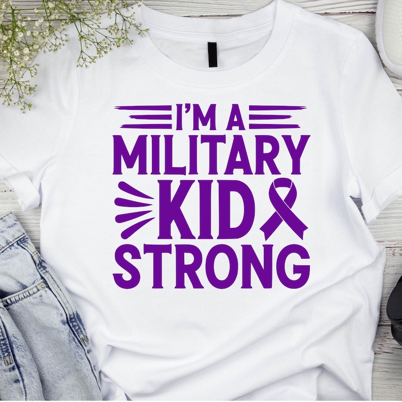 May include: White t-shirt with the purple text "I'M A MILITARY KID STRONG." The design includes a ribbon and stylized text. The shirt is made of a soft material.