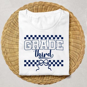 Op de afbeelding: Wit T-shirt met de tekst "GRADE third" in marineblauw. Het woord "GRADE" is in een vetgedrukte, blokletter, en "third" is cursief. Het ontwerp bevat marineblauwe geblokte patronen boven en onder de tekst, en een strik-afbeelding.
