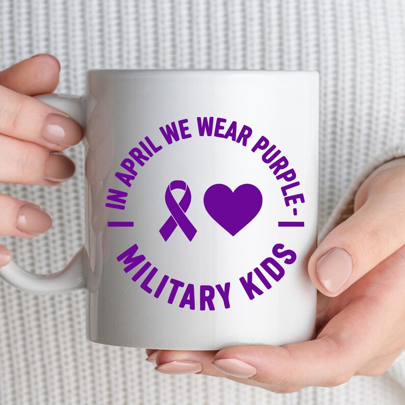 May include: White ceramic mug with the text "IN APRIL WE WEAR PURPLE - MILITARY KIDS" in purple. The design includes a purple ribbon and a purple heart. The mug is held by a person.