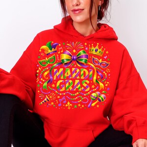 May include: A red hoodie featuring a colorful Mardi Gras design. The graphic includes a bow, crown, masks, musical notes, and the words "MARDI GRAS" in a vibrant, multi-colored font. The design is festive and eye-catching.