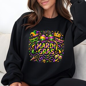 May include: Black sweatshirt featuring a colorful Mardi Gras design. The graphic includes a large purple and gold bow, masks, a crown, musical notes, and the words "MARDI GRAS" in green and yellow. Beads and confetti complete the festive look.