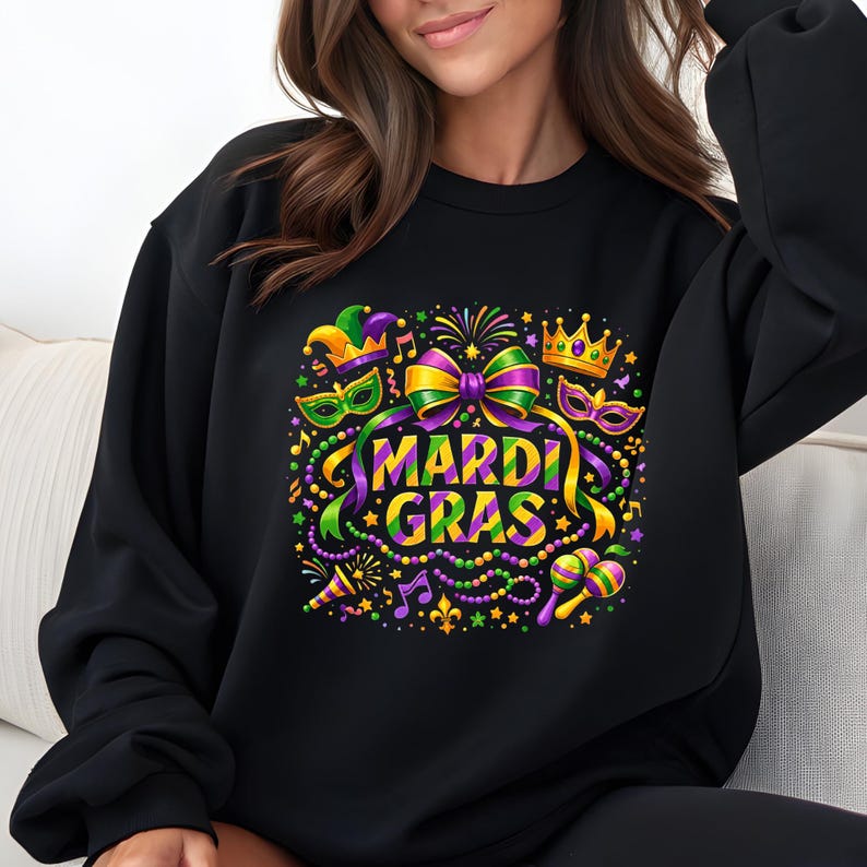 May include: Black sweatshirt featuring a colorful Mardi Gras design. The graphic includes a large purple and gold bow, masks, a crown, musical notes, and the words "MARDI GRAS" in green and yellow. Beads and confetti complete the festive look.