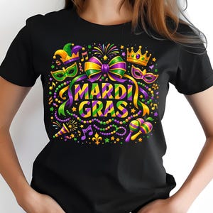 May include: Black t-shirt featuring a colorful Mardi Gras design. The graphic includes a bow, crown, mask, musical notes, and the words "MARDI GRAS" in yellow and green. The design is surrounded by beads and stars.