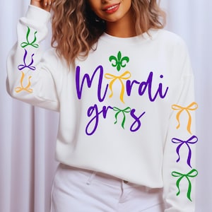Pode incluir: Camisola branca com as palavras "Mardi Gras" em escrita roxa, uma flor-de-lis verde e laços coloridos. As mangas apresentam laços verdes, roxos e amarelos. Uma peça festiva para a época do Mardi Gras.