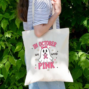 May include: Cream-colored tote bag with the text "IN OCTOBER we WEAR PINK" in pink. A ghost graphic with a pink bow and ribbon is in the center. The bag has a simple design with a light-colored handle.