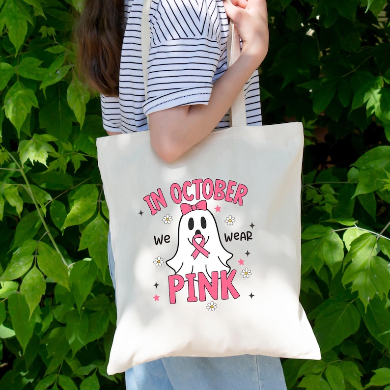 May include: Cream-colored tote bag with the text "IN OCTOBER we WEAR PINK" in pink. A ghost graphic with a pink bow and ribbon is in the center. The bag has a simple design with a light-colored handle.