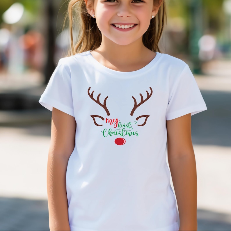 May include: A white t-shirt featuring a reindeer face design with brown antlers, brown eyes, and a red nose. The text "my first Christmas" is written in green and red below the reindeer face. The shirt is worn by a young person.